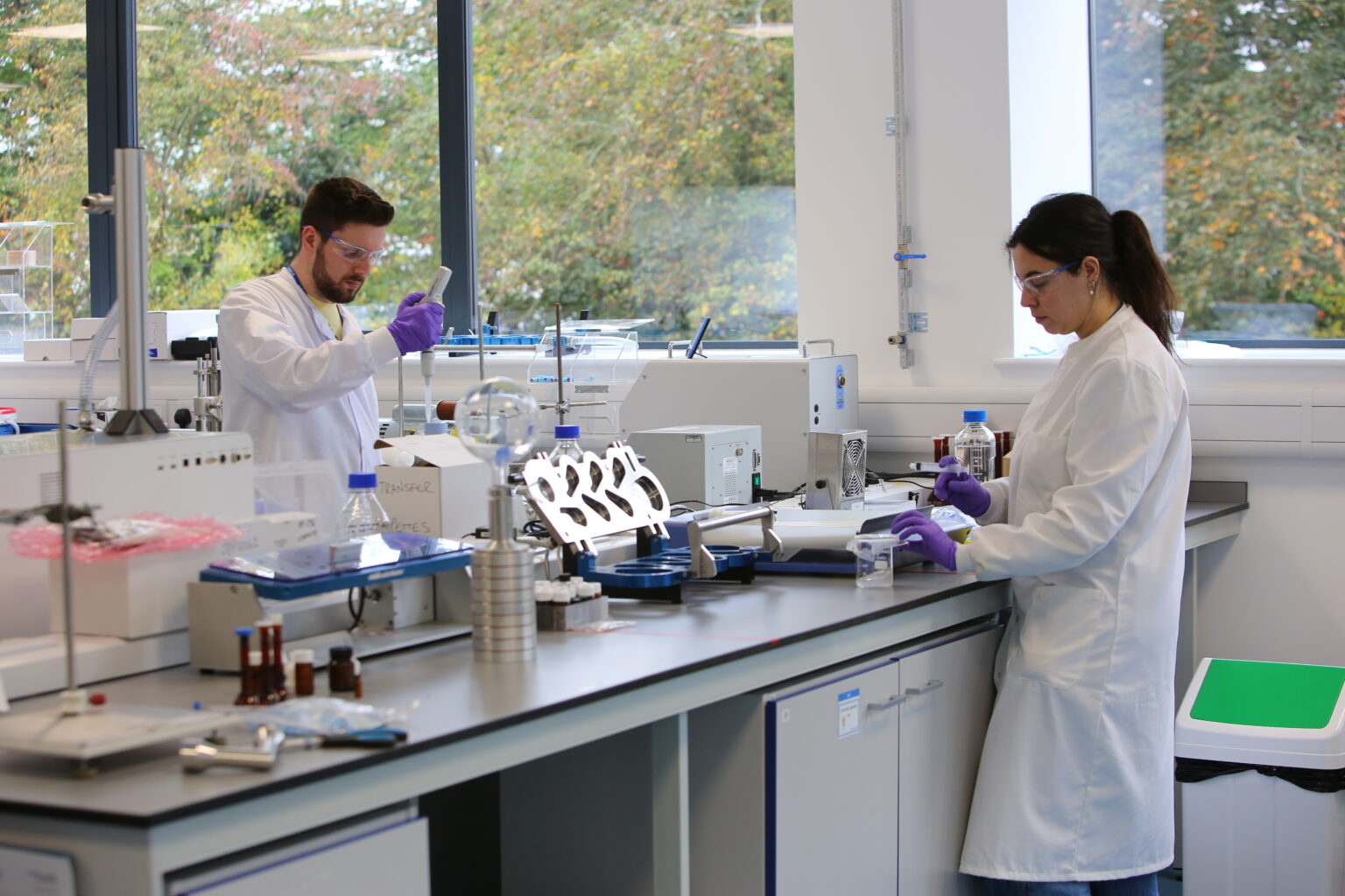 Wide view of windowed cGMP laboratory at Nanopharm with metal APSD test on the black benchtop with two scientists visible.