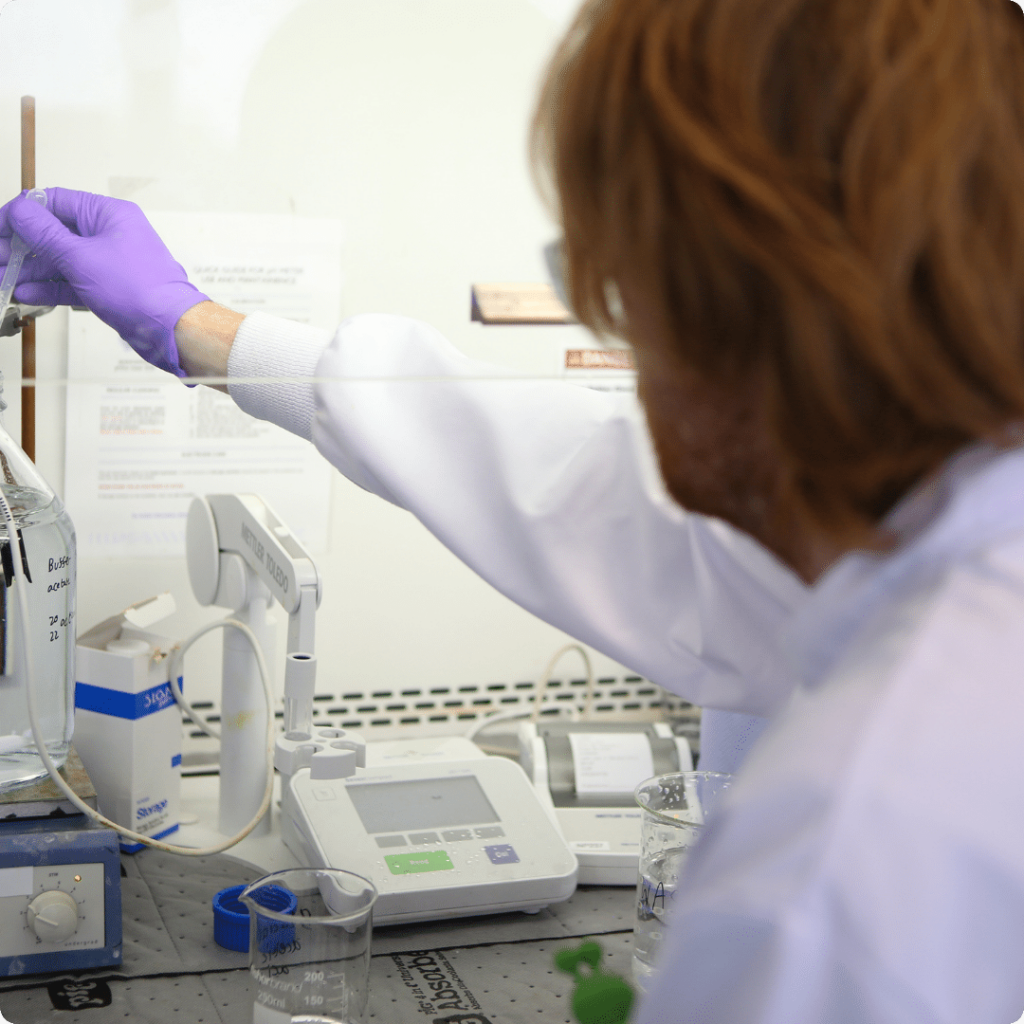 Man with long red hair and white lab coat measures pH of a solution on a stir plate in Nanopharm analytical R&D laboratory.